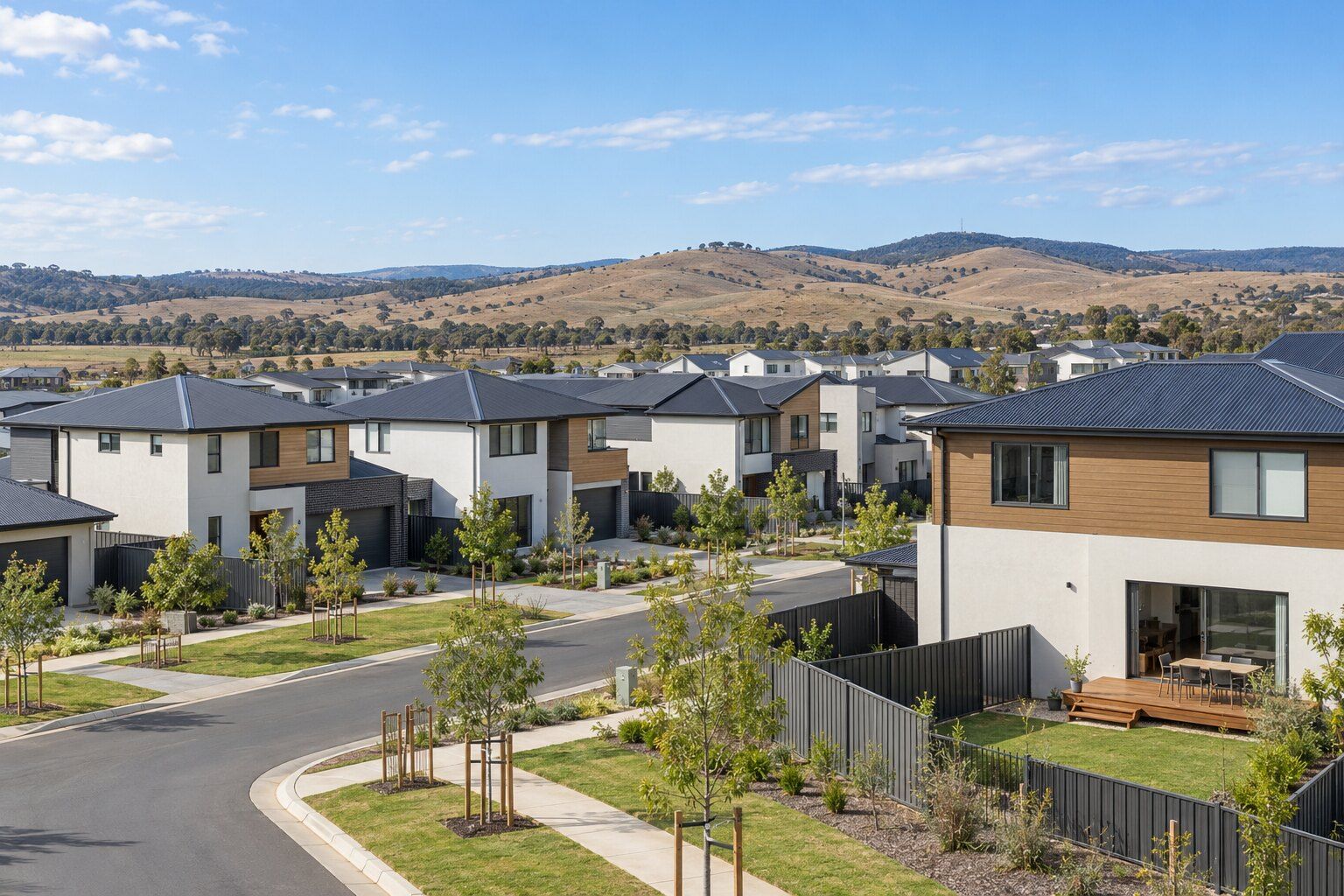 Carpenter, Molonglo Valley: new two-storey homes with timber-look cladding and young staked street trees, one with a low timber deck out the back, and dry grassy hills beyond
