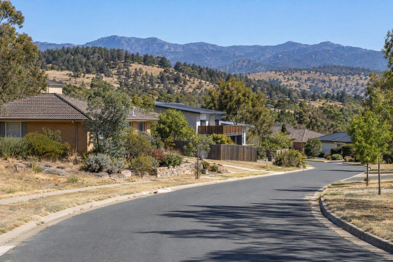 Carpenter, Weston Creek: curving street of brick and rendered houses with a timber balcony and a paling fence, pine-covered hills and a rocky mountain range behind